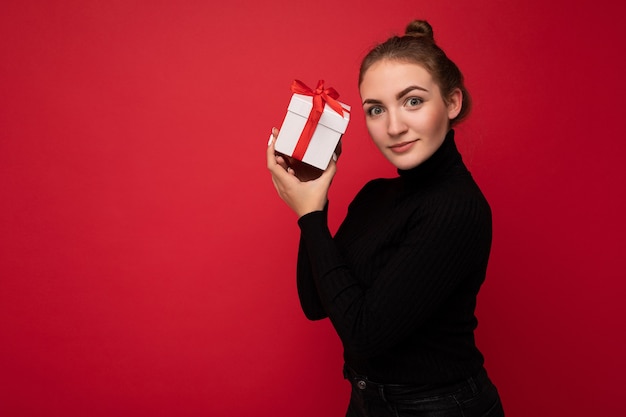 Femme isolée sur mur rouge portant un pull noir tenant une boîte-cadeau blanche avec ruban rouge