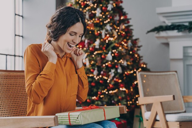 Femme italienne excitée assise sur un canapé près de l'arbre de Noël à la maison avec une boîte-cadeau de Noël emballée