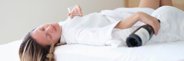 Une femme ivre en peignoir dort sur le lit avec une bouteille de vin et un verre dans la chambre d'hôtel