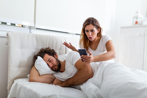 Photo femme jalouse espionnant le téléphone de son partenaire alors qu'il dort dans un lit à la maison. femme jalouse choquée espionnant le téléphone de son mari pendant que l'homme dort dans son lit à la maison