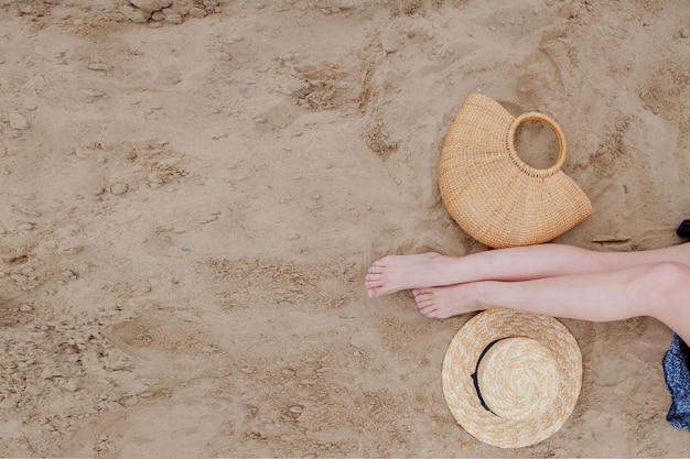 Femme jambes bronzées chapeau de paille et sac sur la plage de sable Concept de voyage Se détendre sur une plage avec les pieds sur le sable