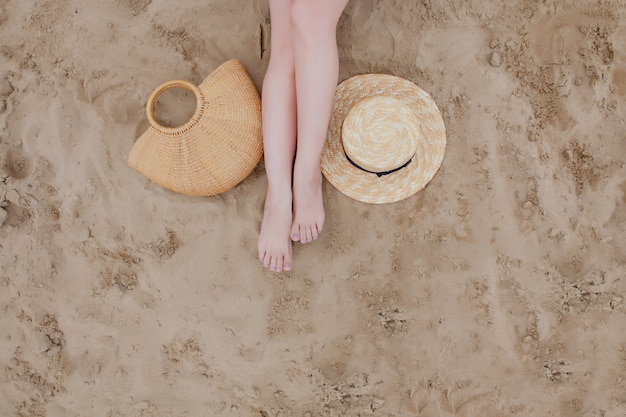 Femme jambes tannées, chapeau de paille et sac sur la plage de sable. Se détendre à la plage, les pieds sur le sable.