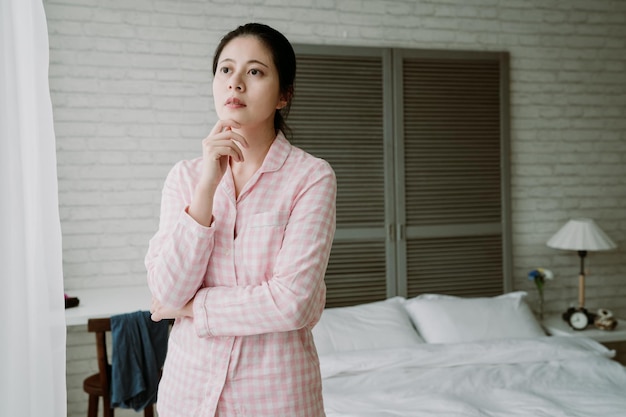 femme japonaise debout à côté de la fenêtre avec un regard pensif. dame asiatique caressant son menton envisage son plan de la journée.