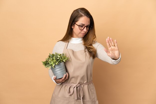 Femme de jardinier d'âge moyen tenant une plante isolée sur beige faisant le geste d'arrêt et déçu