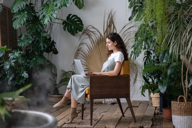 Femme jardinier assis dans un fauteuil en serre travaillant sur un ordinateur portable entouré de plantes d'intérieur exotiques