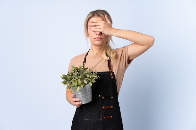 Femme de jardinier blonde tenant une plante sur fond isolé couvrant les yeux par les mains
