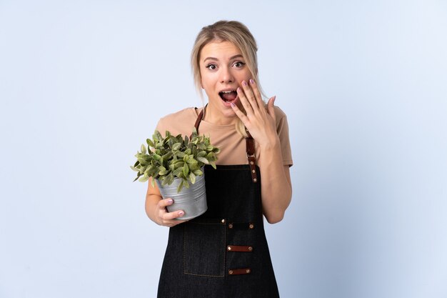 Femme de jardinier blonde tenant une plante sur fond isolé avec une expression faciale surprise