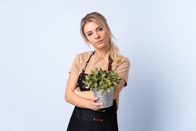 Femme de jardinier blonde tenant une plante sur fond isolé en gardant les bras croisés