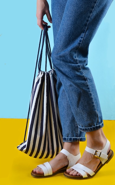 Une femme avec un jean et des sandales tient un sac de plage rayé sur un mur bleu. Heure d'été dans une station balnéaire