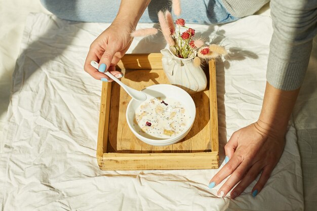 Femme en jeans assise sur le lit et mangeant un bol de granola sain pendant la lumière du soleil du matin, petit-déjeuner au lit.