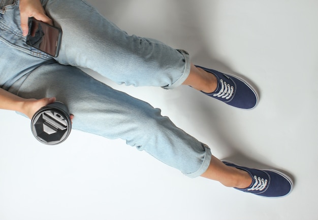 La femme en jeans et baskets est assise sur smartphone et tient une tasse de café. Vue de dessus.