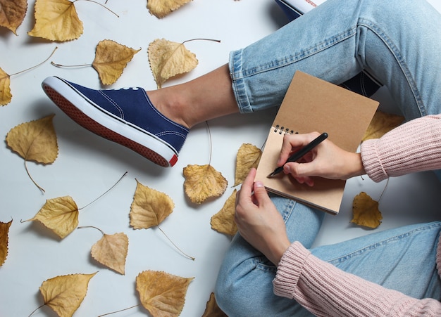 Une femme en jeans et baskets est assise sur un tableau blanc parmi les feuilles jaunes tombées et écrit dans un cahier. Journaliste de travail. Temps de l'automne. Espace de travail.