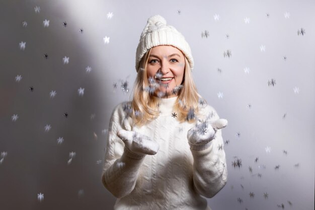 Une femme jette des flocons de neige vêtue d'un chapeau et d'un pull souriant joyeusement.