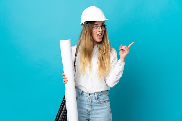 Femme jeune architecte avec casque et tenant des plans isolés sur l'espace bleu pensant une idée pointant le doigt vers le haut