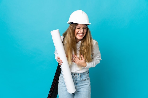 Femme jeune architecte avec casque et tenant des plans isolés sur l'espace bleu souriant beaucoup