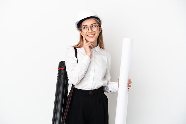 Femme jeune architecte avec casque et tenant des plans isolés sur fond blanc en pensant à une idée tout en levant les yeux