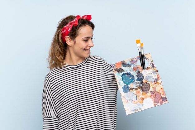 Femme jeune artiste tenant une palette sur un mur bleu isolé avec une expression heureuse