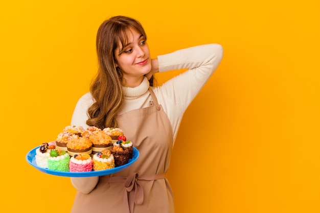 Femme jeune chef pâtissier isolée sur jaune touchant l'arrière de la tête, pensant et faisant un choix.