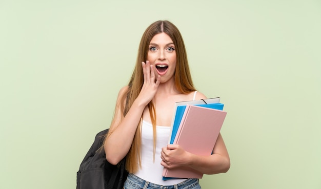 Femme jeune étudiant sur un mur vert isolé avec une expression faciale surprise