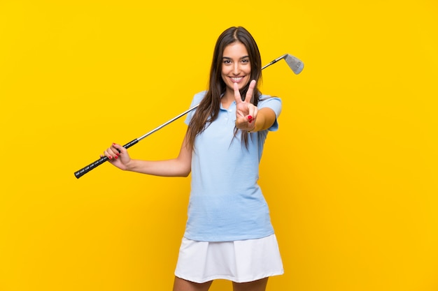 Femme jeune golfeur sur mur jaune isolé, souriant et montrant le signe de la victoire