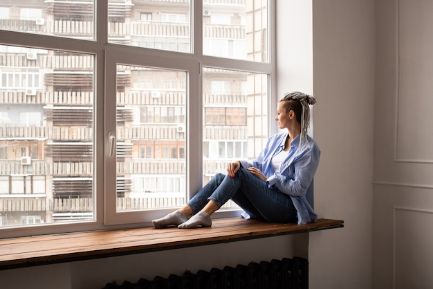 Femme jeune hipster est assise sur le rebord de la fenêtre. Seul à la maison en quarantaine. Thème du coronavirus. Reste à la maison