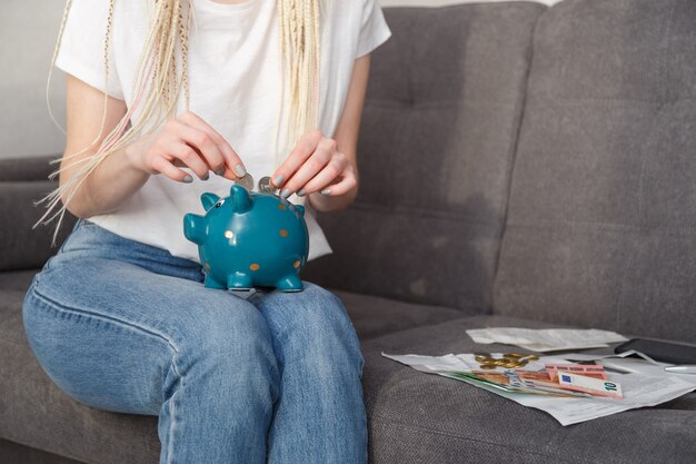 Femme jeune hipster mettant des pièces dans une tirelire pour voyager assis sur un canapé à la maison. Concept d'épargne