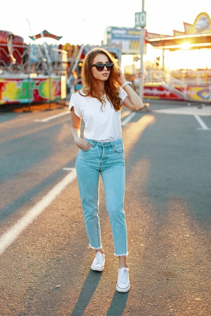 Femme jeune hipster en T-shirt élégant dans des lunettes de soleil à la mode en blue-jeans en baskets posant dans un parc d'attractions