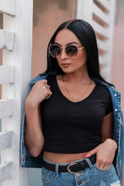 Femme jeune hipster urbaine moderne dans des lunettes de soleil noires élégantes se dresse et redresse une veste bleu denim près d'un mur en bois vintage. Modèle de jolie fille brune dans des vêtements à la mode posant à l'extérieur.