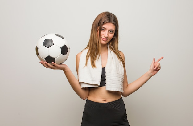 Femme jeune de remise en forme pointant vers le côté avec le doigt et tenant un ballon de soccer.