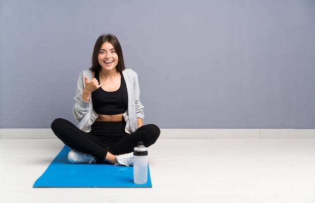 Femme jeune sport assis sur le sol avec tapis faisant un geste de téléphone