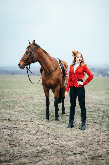 Femme jockey aux cheveux roux dans un cardigan rouge et des bottes noires avec un cheval pour une promenade