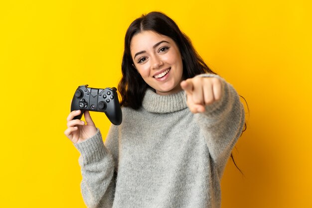 Femme jouant avec un contrôleur de jeu vidéo isolé sur mur jaune pointe du doigt vous avec une expression confiante