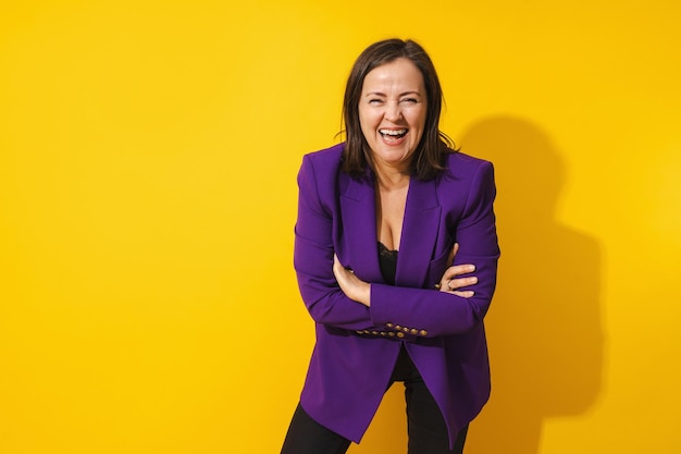 Une femme joyeuse d'âge moyen portant un blazer violet souriant sur un fond jaune