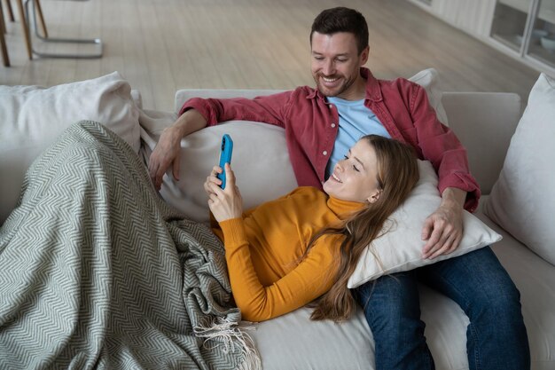 Une femme joyeuse allongée sur les genoux d'un homme tenant un smartphone souriante tout en faisant un appel vidéo avec des parents