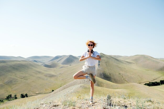 Femme joyeuse aux cheveux bouclés debout sur une colline, en équilibre sur un pied. Posant pour une photo, souriant à la caméra. De belles collines qui s'étendent partout.