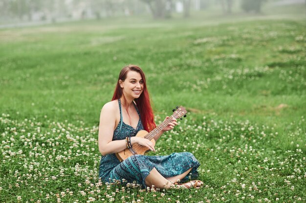 Femme joyeuse aux cheveux rouges jouant du ukulélé dans le parc assis sur l'herbe un jour d'été.