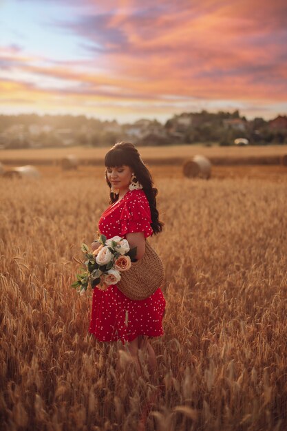 Femme joyeuse aux longs cheveux noirs ondulés en robe rouge avec un sac et des fleurs dans ses mains