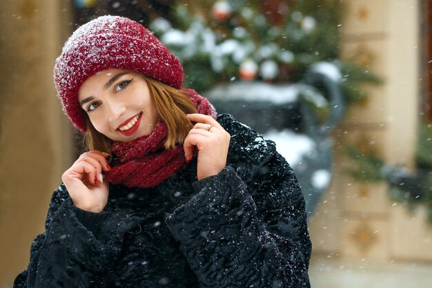 Femme joyeuse brune aux lèvres rouges portant une casquette et une écharpe rouges marchant dans la ville pendant les chutes de neige. Espace libre