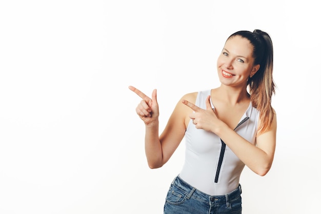 Photo une femme joyeuse dans un haut blanc sans manches et un jean denim pointe vers son côté avec les deux mains son sourire ludique suggérant quelque chose d'excitant hors cadre sur un fond blanc