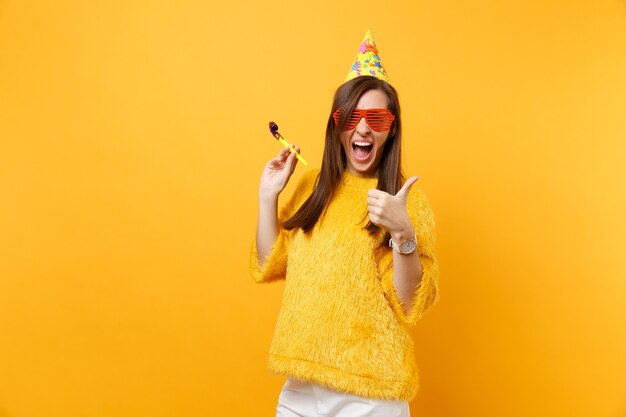 Femme joyeuse dans des lunettes oranges drôles, chapeau d'anniversaire avec pipe jouant montrant le pouce vers le haut, célébrant isolé sur fond jaune vif. Concept de mode de vie des émotions sincères des gens. Espace publicitaire.