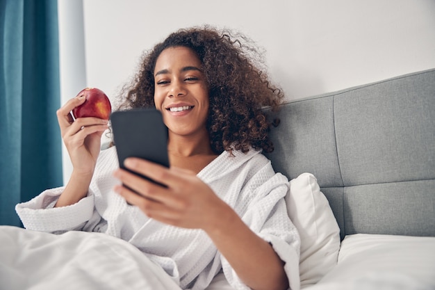 Femme joyeuse dans un peignoir gaufré tenant un téléphone portable et une pomme dans ses mains