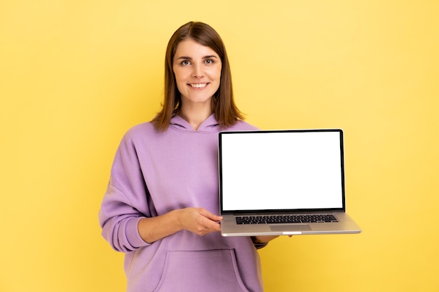 Femme joyeuse debout montrant un ordinateur portable avec un écran blanc vide pour la promotion, regardant la caméra avec le sourire, portant un sweat à capuche violet. Studio intérieur tourné isolé sur fond jaune.