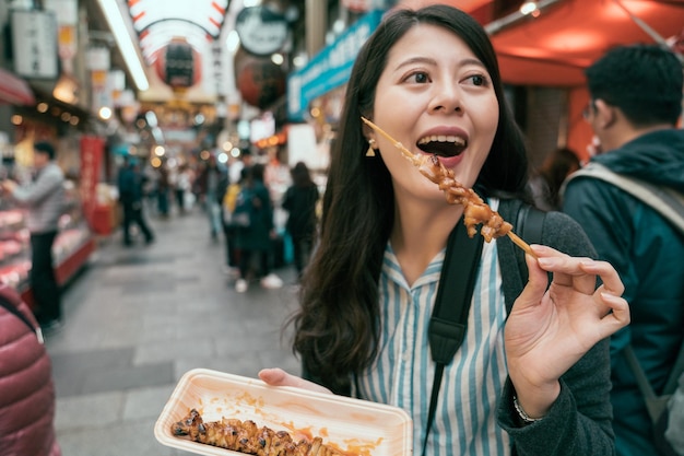 une femme joyeuse découvre la culture japonaise locale en mangeant de savoureux yakitori. joyeux touriste mangeant dans un marché couvert au Japon. marché grouillant à osaka.