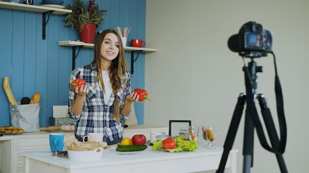 Femme joyeuse enregistrant un blog vidéo sur des aliments sains végétariens sur un appareil photo reflex numérique dans la cuisine à la maison