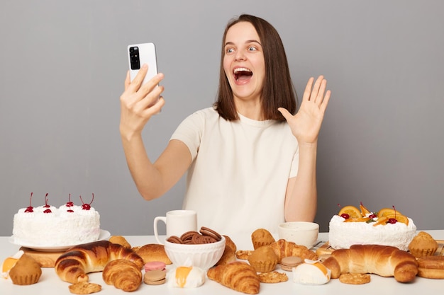 Femme joyeuse étonnée portant un t-shirt blanc isolé sur fond gris faisant selfie ou diffusant en direct en agitant la main en disant bonjour aux abonnés