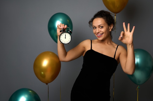 Une femme joyeuse et heureuse de race mixte tient un réveil, il est minuit, montrant des sourires de signe OK regardant la caméra, isolée sur fond gris avec des ballons. Nouvel an, concept de joyeux Noël pour la publicité