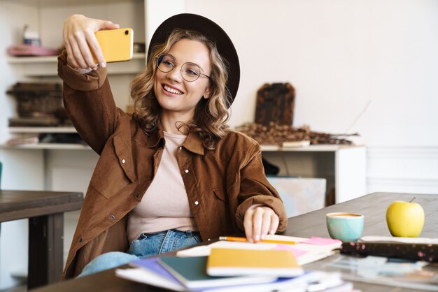 femme joyeuse à lunettes et chapeau prenant selfie sur téléphone portable tout en étudiant avec des cahiers à la maison