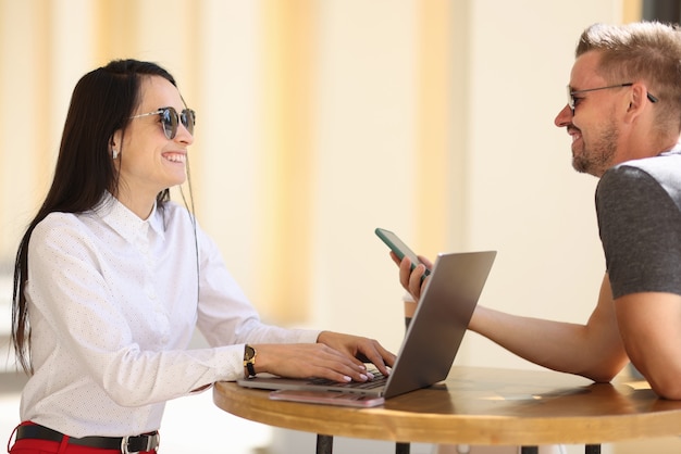 Femme joyeuse à lunettes de soleil s'asseoir à table ronde dans la rue et tapez sur ordinateur portable. L'homme rencontre la femme et prend le numéro de téléphone.