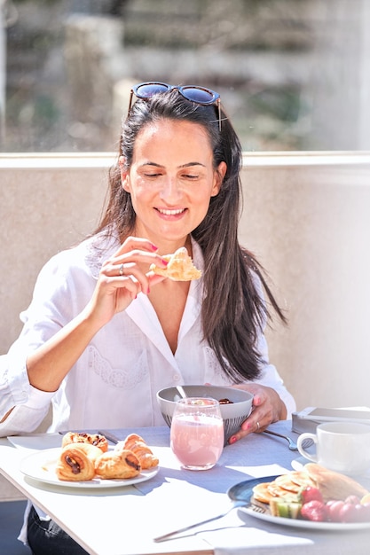 Une femme joyeuse mangeant un délicieux croissant avec du porridge tout en prenant le petit déjeuner dans la véranda le matin d'été