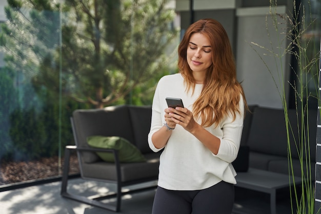 Femme Joyeuse Passant Du Temps à La Maison Et Souriant Tout En Regardant Le Téléphone Et En écrivant Un Message à Quelqu'un. Concept De Personnes Et De Technologies